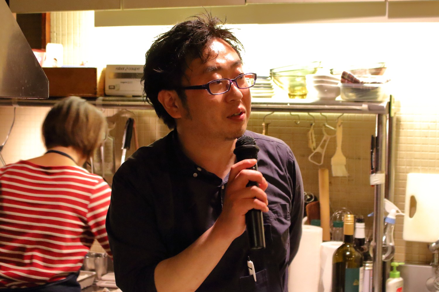 背景では別の人物が料理をしている様子が見えるキッチンの前で、紺色のシャツを着た店主の男性がマイクを持って話している写真