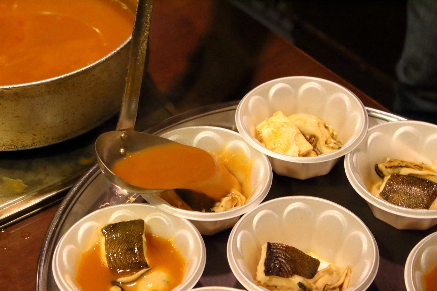 お盆に牡蠣と白身魚が入った使い捨てカップが並び、鍋からお玉でオレンジ色のスープをすくって注いでいるブイヤベースの料理の写真
