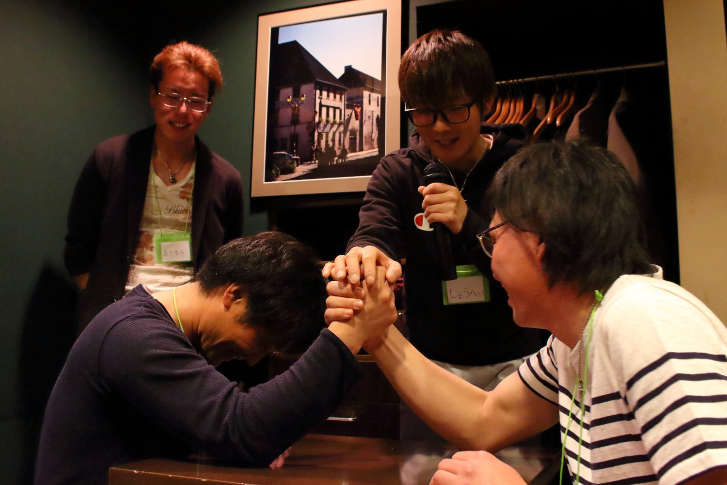 周囲で男性二人が応援や実況をしている室内のテーブルの上で、二人の男性が腕相撲をしている腕相撲大会の様子の写真