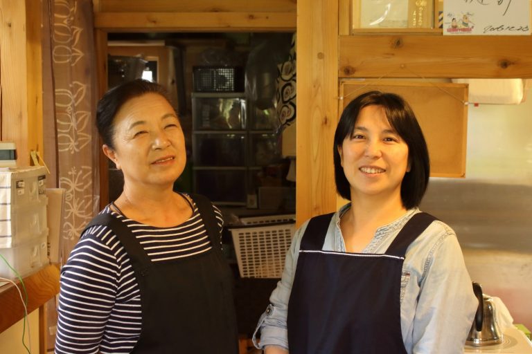 サインが飾られたカフェレストランの店内で、黒いエプロンを着用した2名の女性が笑顔で並んで写っている写真