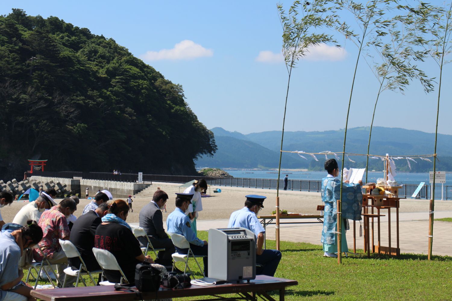 青空の下、海岸近くの芝生の上で、和服の男性が竹で囲まれた祭壇の前に立ち、スーツや制服を着た人々が椅子に座り安全祈願祭が行われている写真