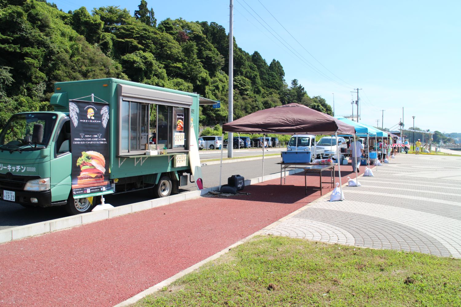 青空の下、緑の木々が茂る道路沿いに緑色の移動販売車が停車し、その隣には複数のテントが並んで設置されているテント販売所の写真