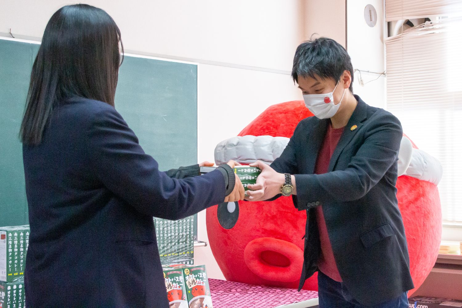 オクトパス君のキャラクターの着ぐるみが見ているなか、スーツ姿の男性が女子高校生にオクトパス君合格カレーの箱を両手で手渡している様子の写真