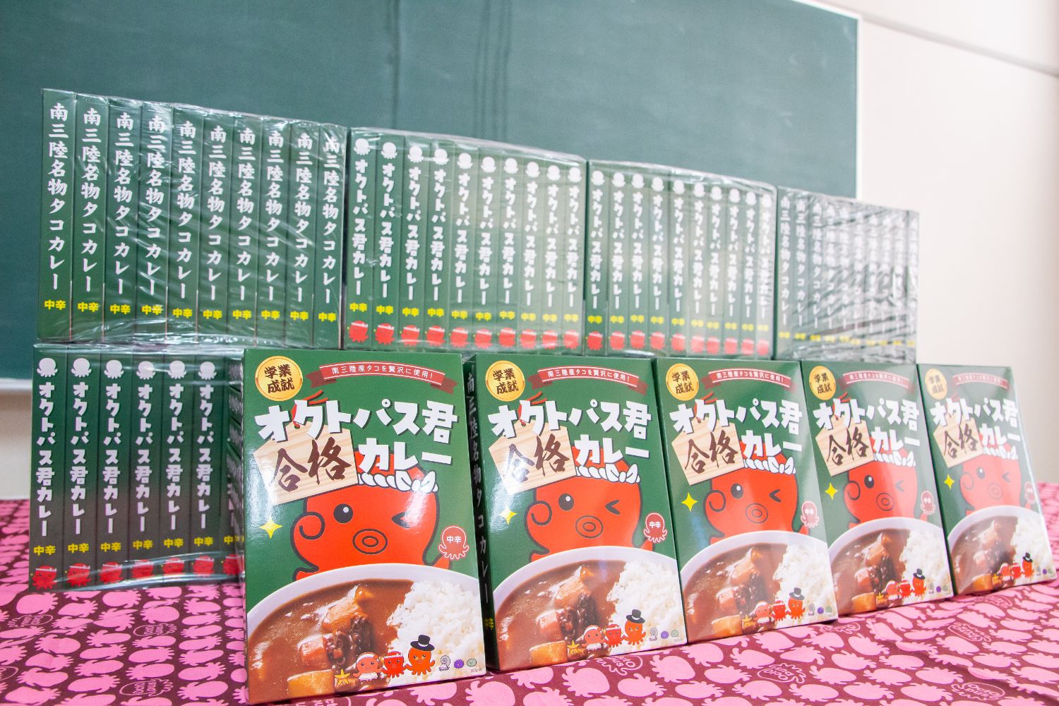 黒板の前の机に、プレゼントされたオクトパス君合格カレーがパッケージの絵柄が見えるように多数積み上げられている写真