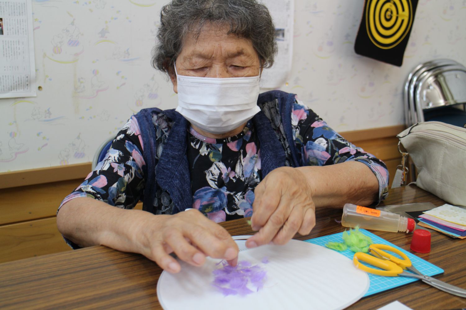 マスクをつけた高齢の女性が、机に置いた白いうちわの上にちぎった薄紫色の和紙を丁寧に貼り付けて装飾している写真