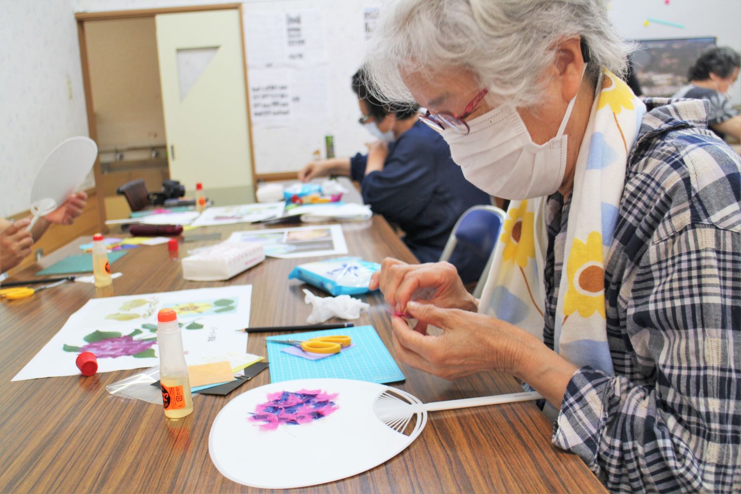 マスクをつけた白髪の女性が真剣な表情で机の上の白いうちわに、ピンクと紫色の和紙をちぎって花の装飾を貼り付ける作業を行っている様子を左から撮影した写真