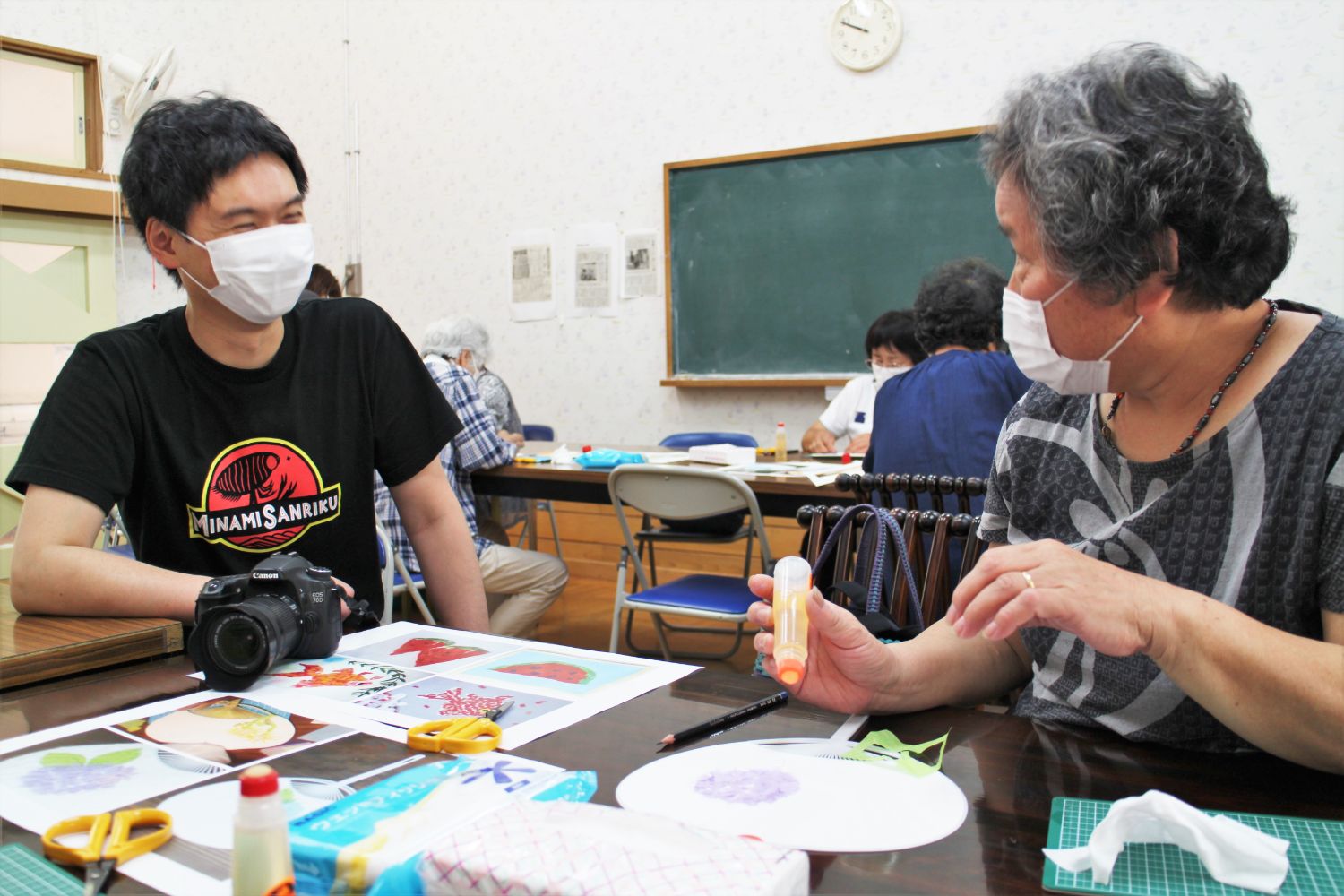 年配の女性がうちわ作りの作業中に液体のりを手にしながら、カメラを置いた隣のテーブルに座る黒いシャツを着た大森さんと笑顔で会話をしている写真