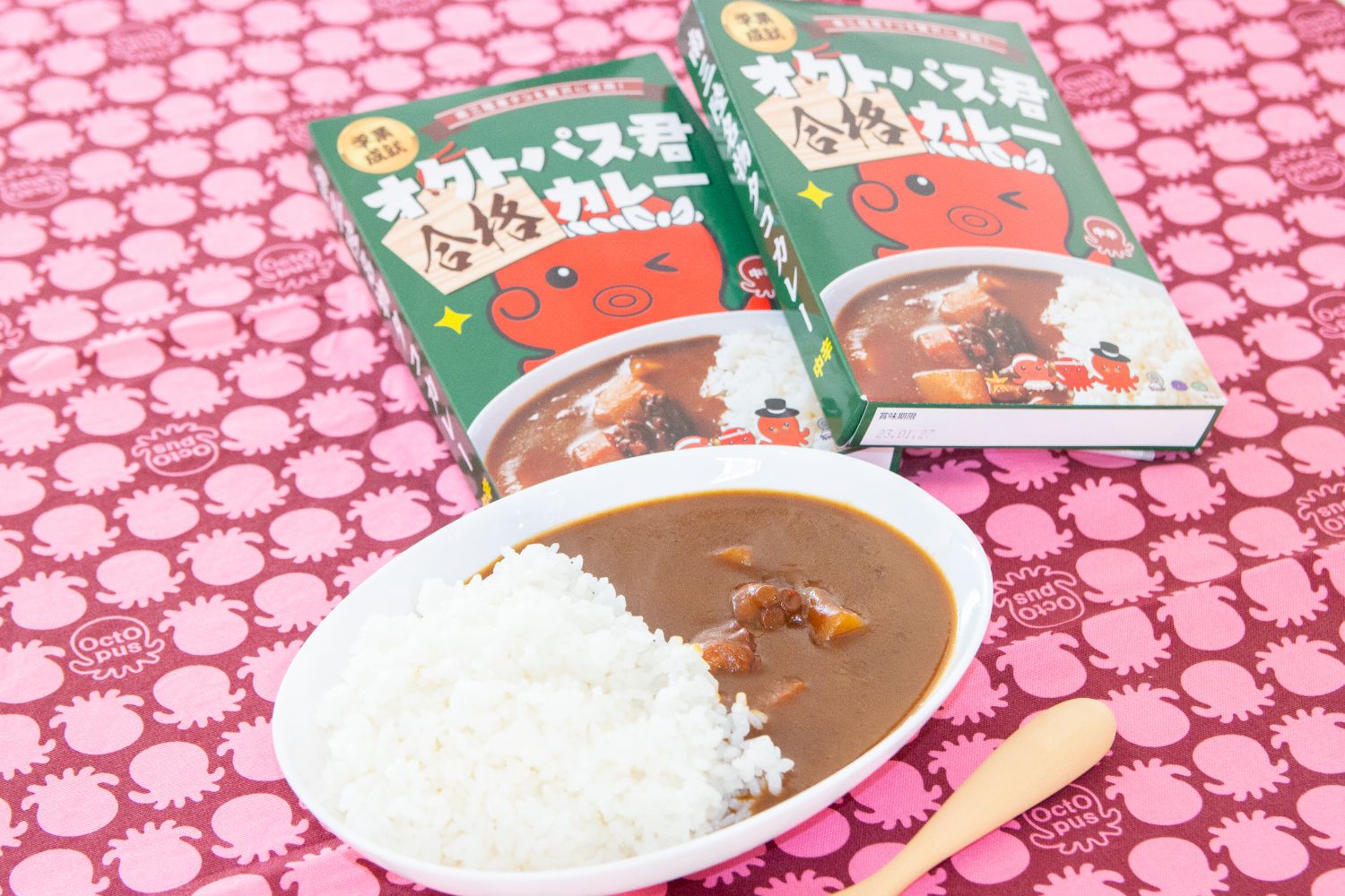 ピンク色のタコ柄クロスの上にオクトパス君合格カレーのパッケージが2つ並べられ、手前のカレー皿にご飯と具入りルーが盛られたレトルトカレーが置かれている写真