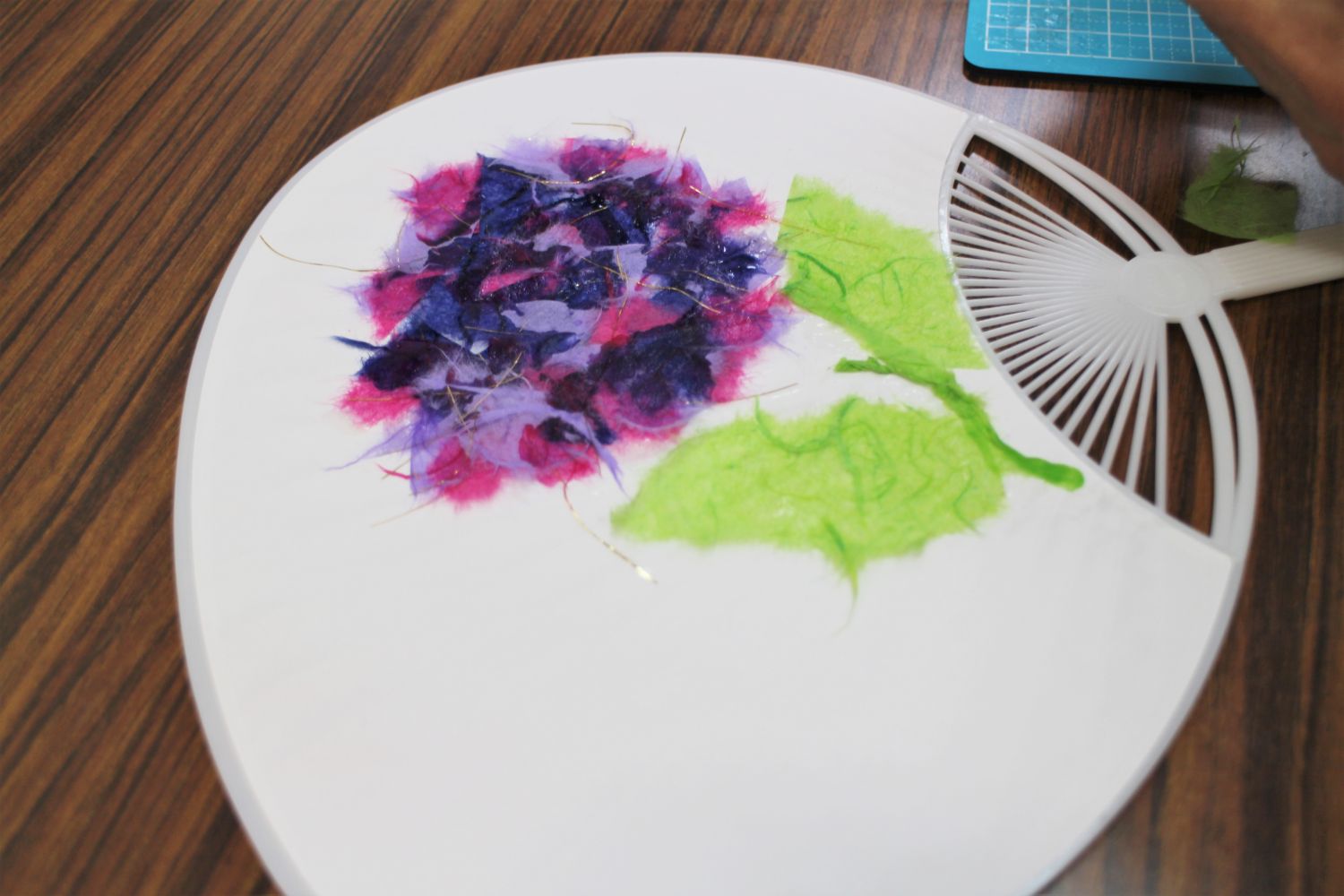 白いうちわの上に紫とピンクのアジサイの花と緑の葉が貼り付けられ、細かな繊維や金の糸があしらわれている手作りうちわの作品の写真