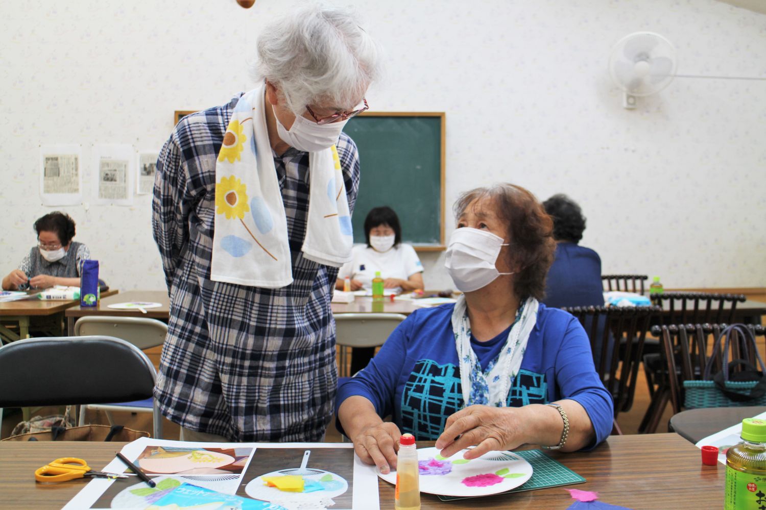 マスクをつけた年配の女性二人が机の前で向かい合い、一人がうちわの制作を進めながらもう一人が立って会話している写真