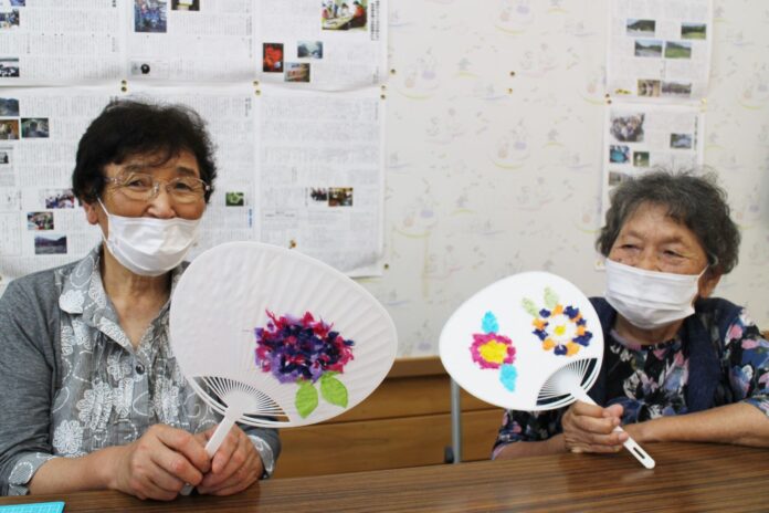 年配の女性二人が笑顔で机に並んで座り、マスクをつけながら、それぞれが手作りの花が飾られた白いうちわを持ってカメラに向かって見せている写真