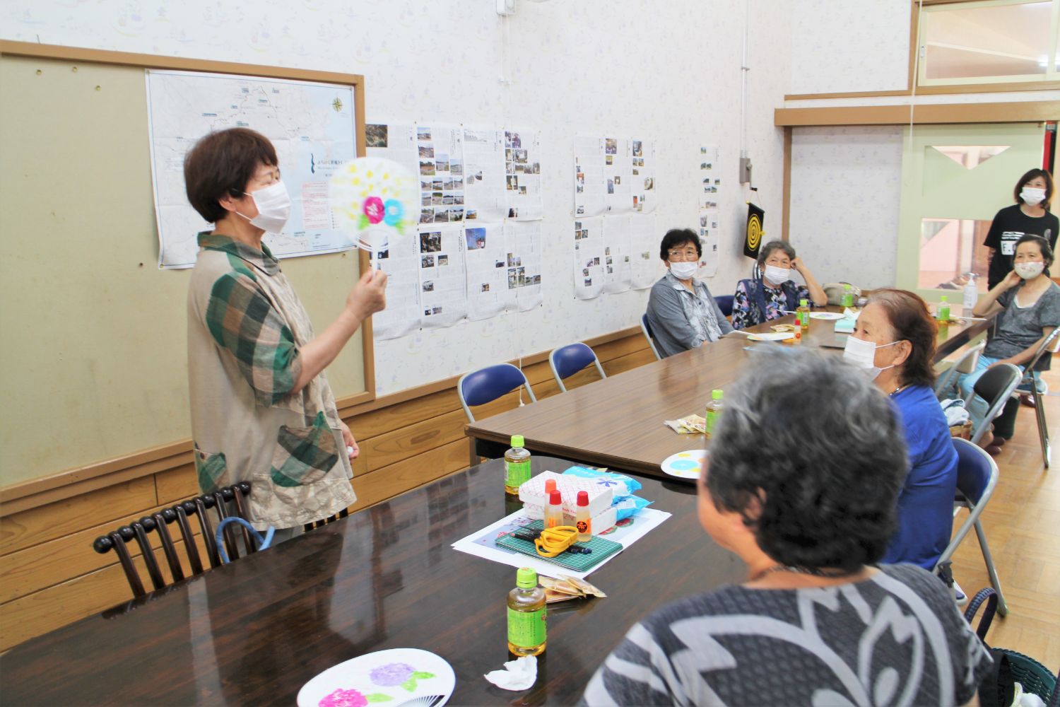 マスクをつけた講師のような女性が完成した装飾うちわを持ち上げて見せ、机の周りに座った年配の参加者たちが注目している写真