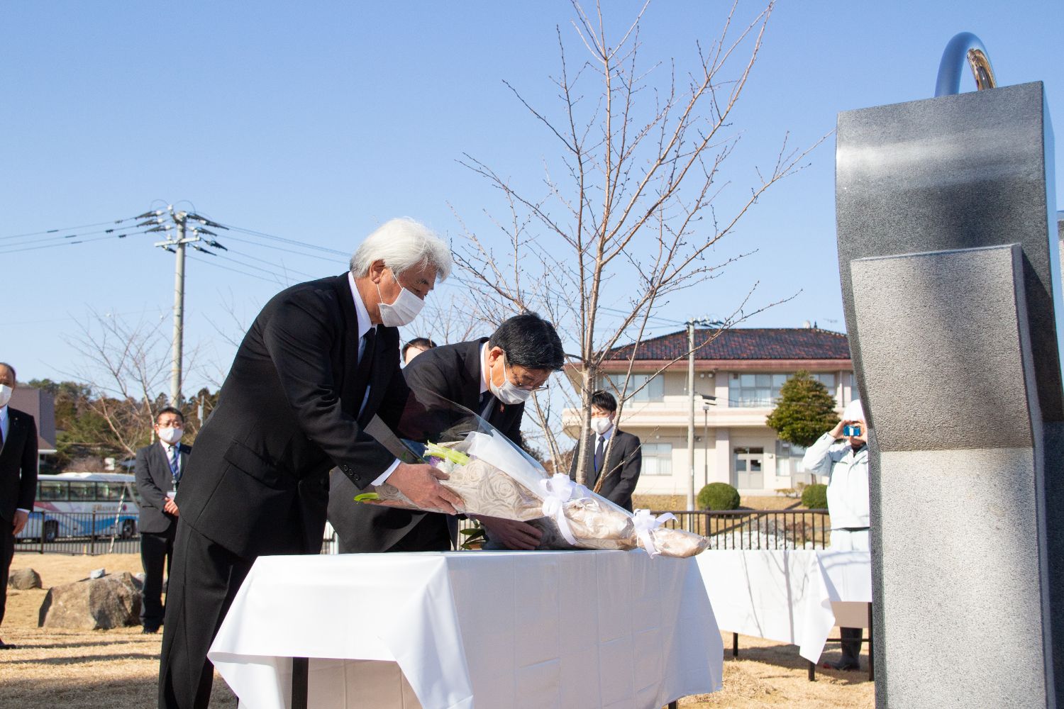 黒いスーツを着た2人の男性が白い布をかけた台の上に白い花束を丁寧に供えている写真