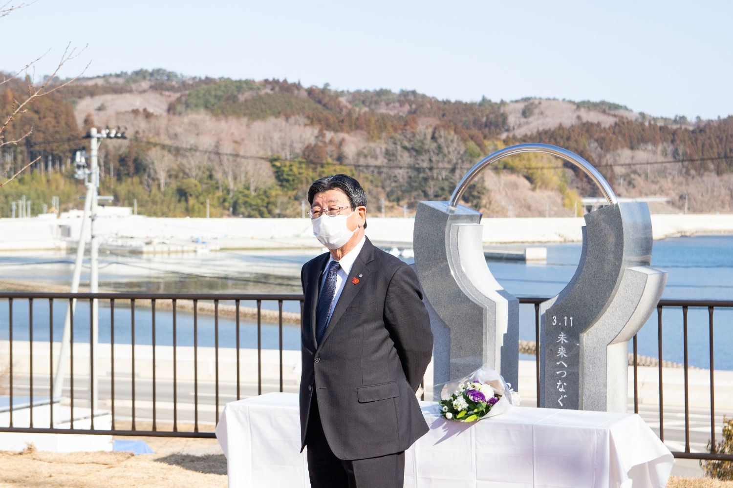 海を背景にした記念モニュメントの前で黒いスーツ姿の男性がマスクを着けて立っており、献花台の上には紫と白の花束が供えられている、追悼の場面を写した写真