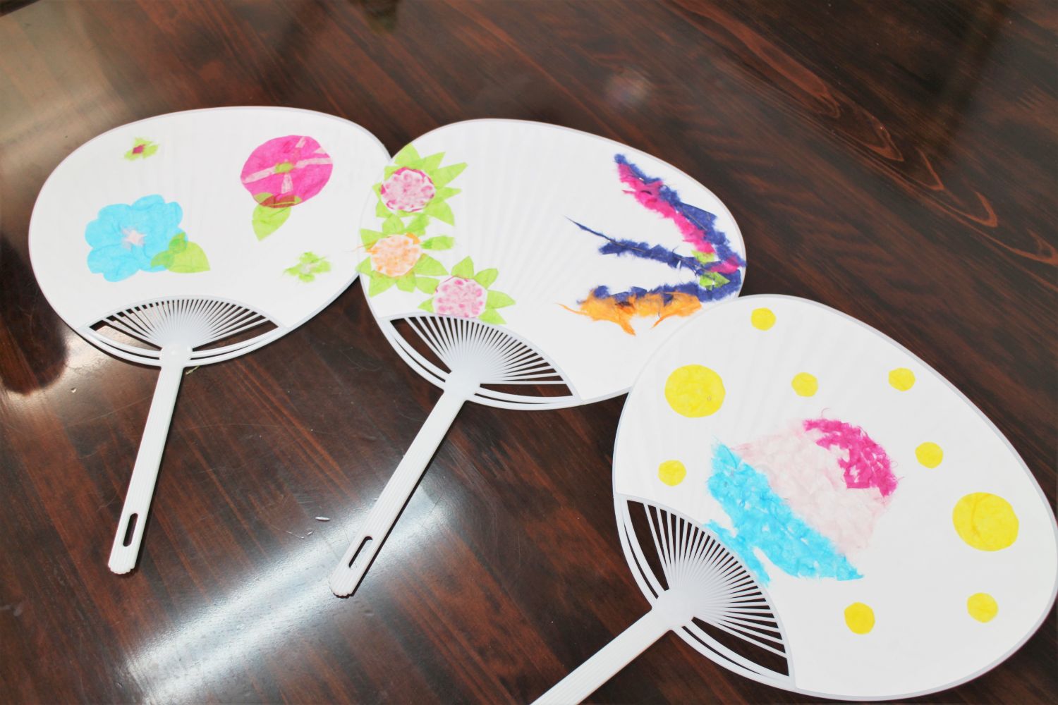 机の上に並べられた三本の白いうちわに、青やピンクのアサガオやかき氷、赤やオレンジ色の花などが貼り付けられた作品が並ぶ写真