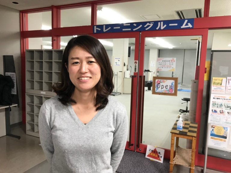 灰色のセーターを着た女性が「トレーニングルーム」と書かれた青い看板のあるガラス扉の前に立っている様子をアップで撮影した写真