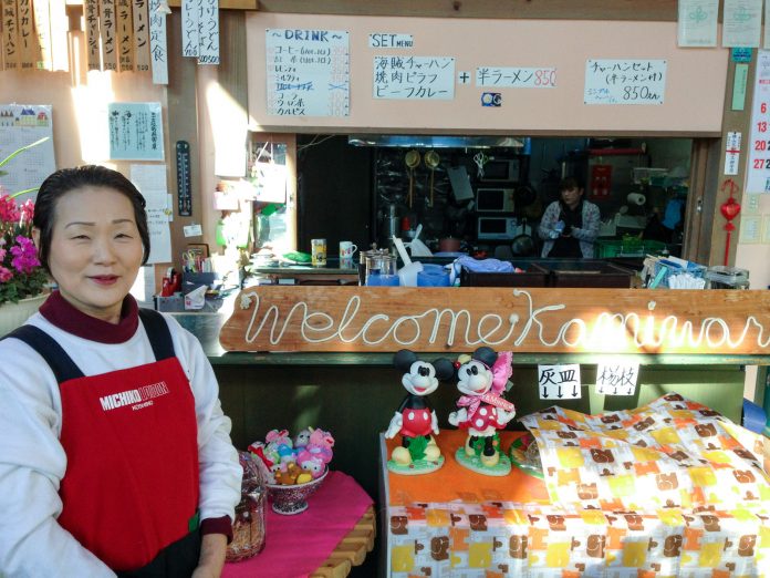 奥の壁には飲み物や食べ物のメニューが貼られ、人形や置物が飾られた店内のカウンター前に赤いエプロン姿の女性が笑顔で立っている写真