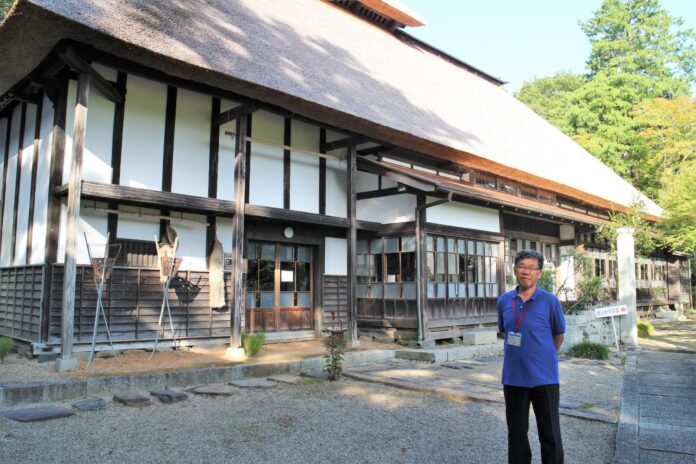 大きな茅葺屋根のひころの里の建物の前で両手を後ろで組み立っている鈴木清美さんの写真