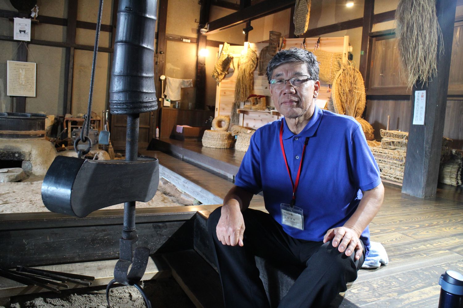 藁で作られた生活用品や民具が棚に並べられている木造の伝統的な和室の中で、青いポロシャツを着て眼鏡をかけた鈴木さんが囲炉裏の横に座っている写真