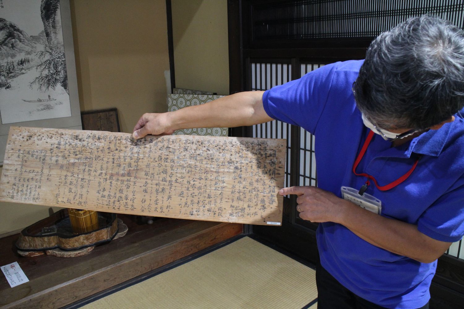 施設所長の鈴木さんが、筆で書かれたたくさんの漢字が並ぶ古い木の板を両手で持ち、その内容を指で示している写真
