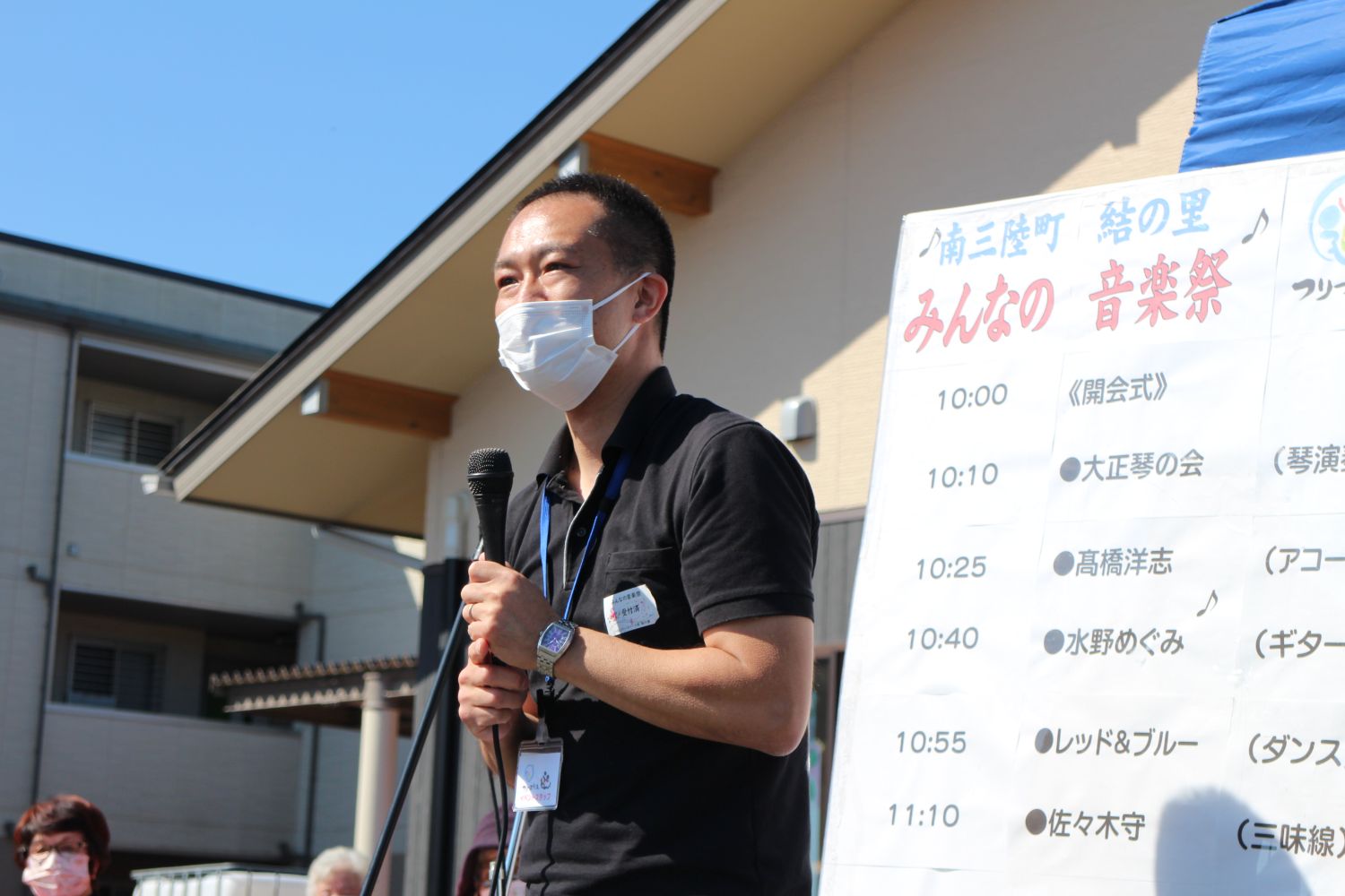 音楽祭のスケジュールが書かれた看板の横で、マイクを手に開会のあいさつをしている高橋雅道さんの写真