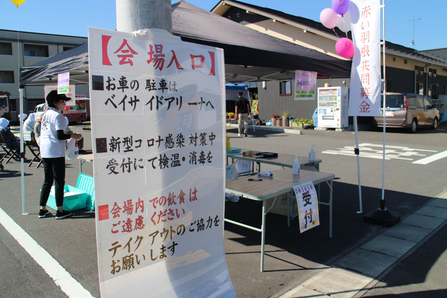 会場入り口に設置された受付の様子で、新型コロナ感染対策の案内や消毒液が置かれた机、赤い羽根共同募金ののぼりが立っている様子の写真