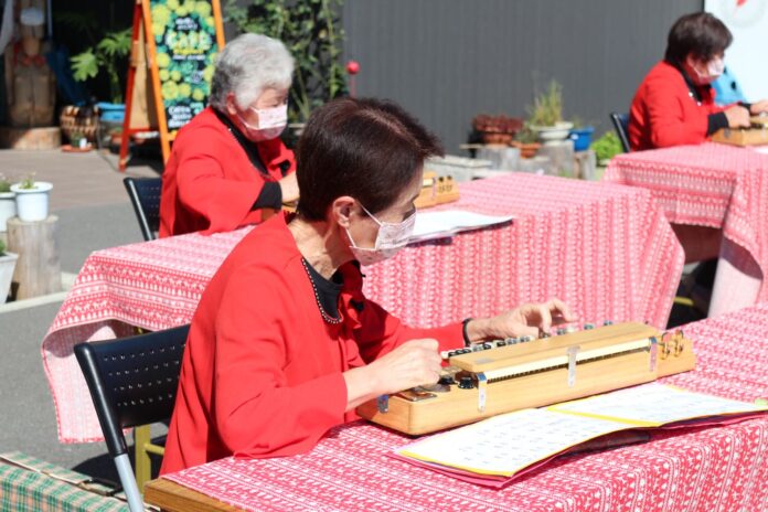 屋外にテーブルが並べられ、テーブルに楽譜を広げ、赤い衣装の高齢女性たちが大正琴を演奏している様子の写真