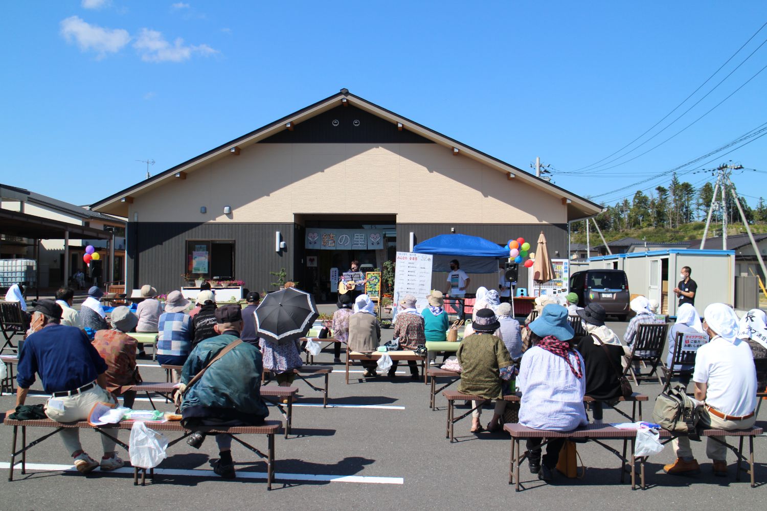 青空の下、地域の『結の里』施設前に設置されたステージ前で、多くの観客がベンチに座り音楽祭を楽しんでいる様子の写真