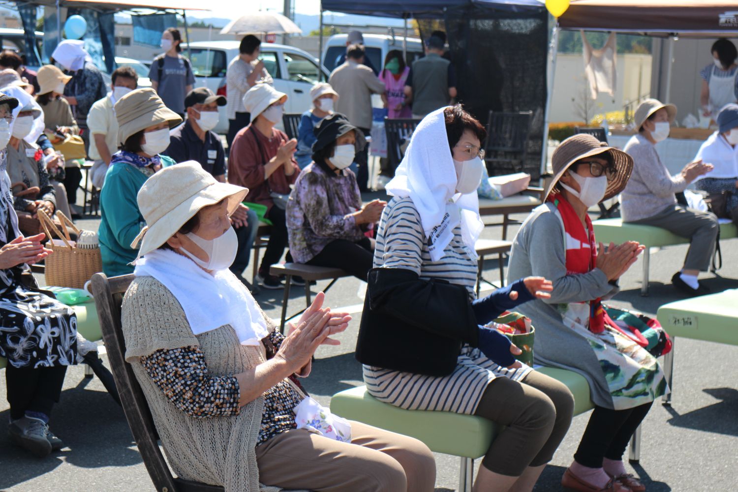 日差しよけにタオルやスカーフを頭や首に巻いている人や、マスクを着用し帽子をかぶった方々が屋外の椅子に座り、前方での発表や演奏を見て拍手をしている様子の写真