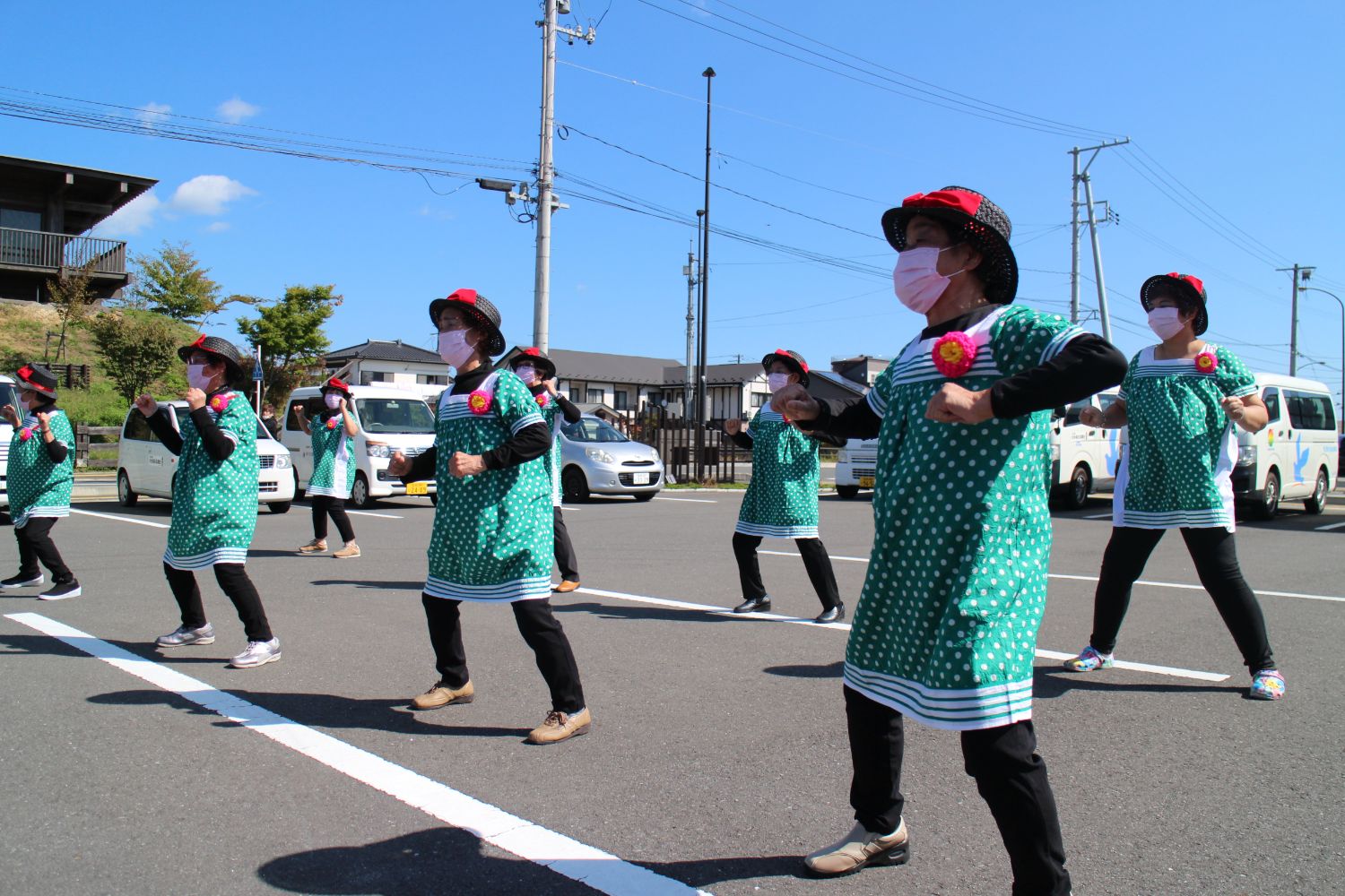 緑の水玉模様の服と黒い帽子を身に着け、ピンクのマスクをして並んで屋外の駐車場で、両腕を前に曲げて拳を握り、足を少し広げて踏み込むような動きのダンスを披露している『レッド&ブルー』のみなさんの写真