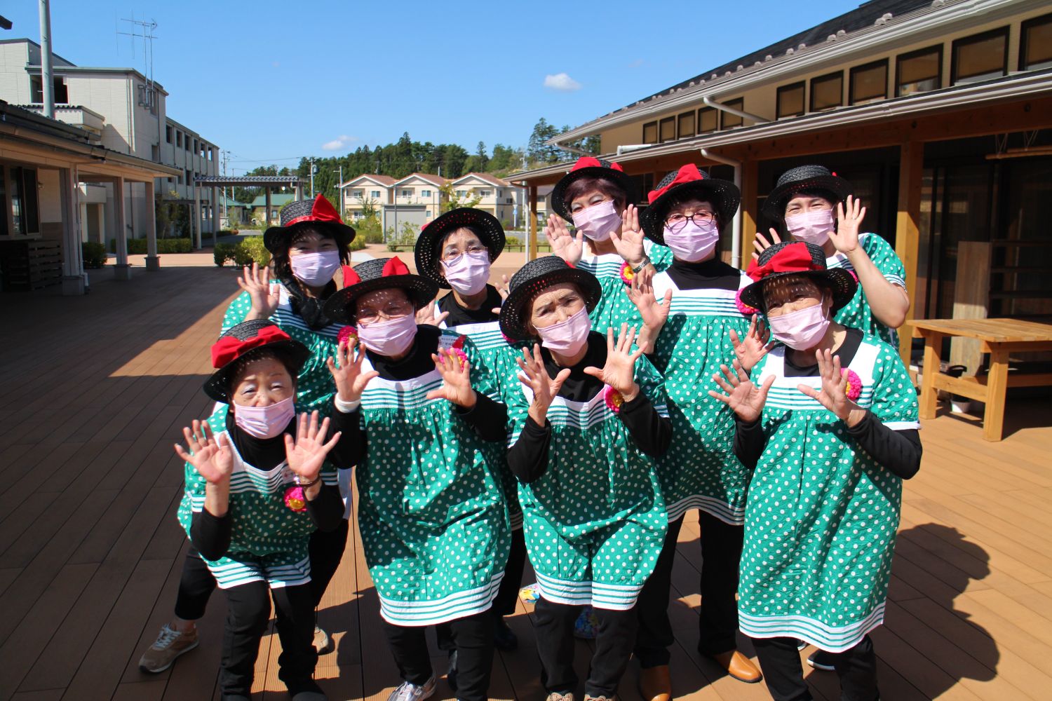 緑の水玉模様の揃いの衣装と黒い帽子をかぶり、ピンクのマスクをつけて両手をあげて笑顔でポーズをとっている女性グループ、レッド&ブルーのみなさんの写真