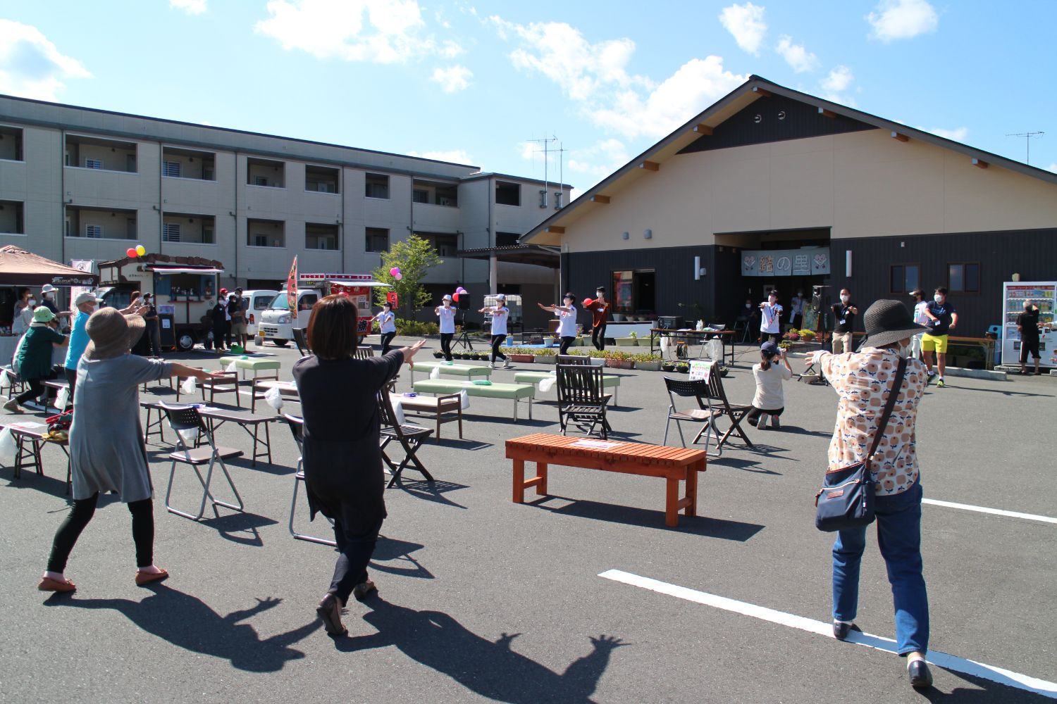 青空の下で広い駐車場に設置された椅子やベンチの前で、多くの人が腕をまっすぐ前に伸ばし、片足を前に出した長生き坂音頭を一緒に踊っている写真