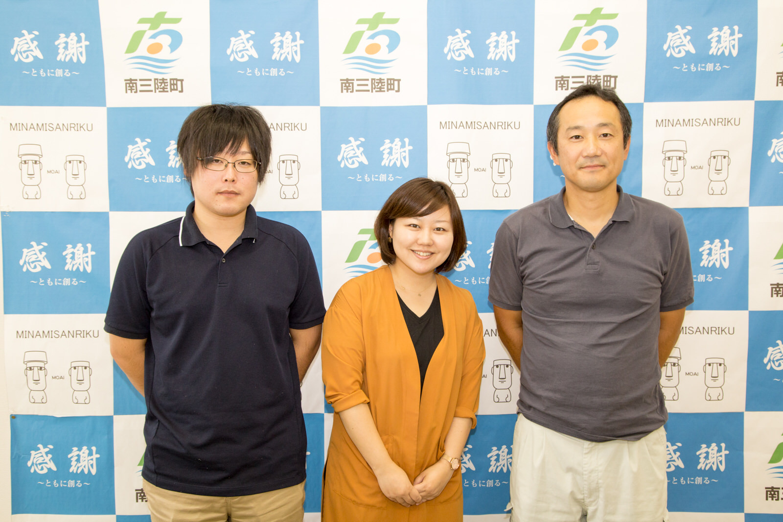 南三陸町のバックパネルの前で、女性リポーターを中央に2人の男性が並んで立っている写真