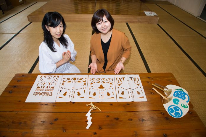広い和室の木の台の上に4枚の切り紙作品と模様が描かれた3つのうちわ、紙垂が並べられ、その前に女性リポーターと白い洋服の工藤さんが並んで座っている写真