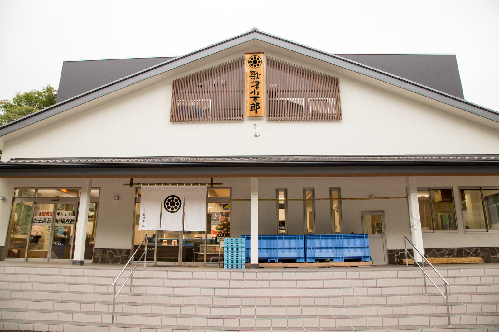 「歌津小太郎」と書かれた木製の看板が掲げられた、白壁と瓦屋根の和風建築の建物の前に、積み重なった青いプラスチックケースが置かれ、入り口前の軒下には紋入りの白いのれんがかかっている店の外観写真