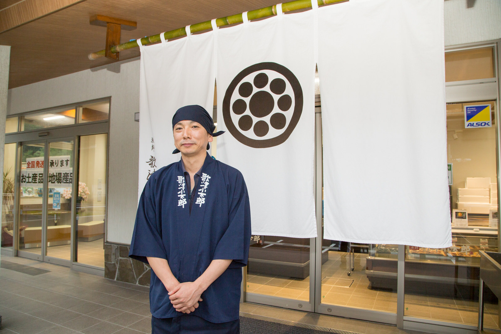 紺色の布を頭に巻き、同じ色の「歌津小太郎」と書かれた法被を身につけた男性が、は丸の中に点を配した紋が描かれた白いのれんの前で、笑顔で立っている写真