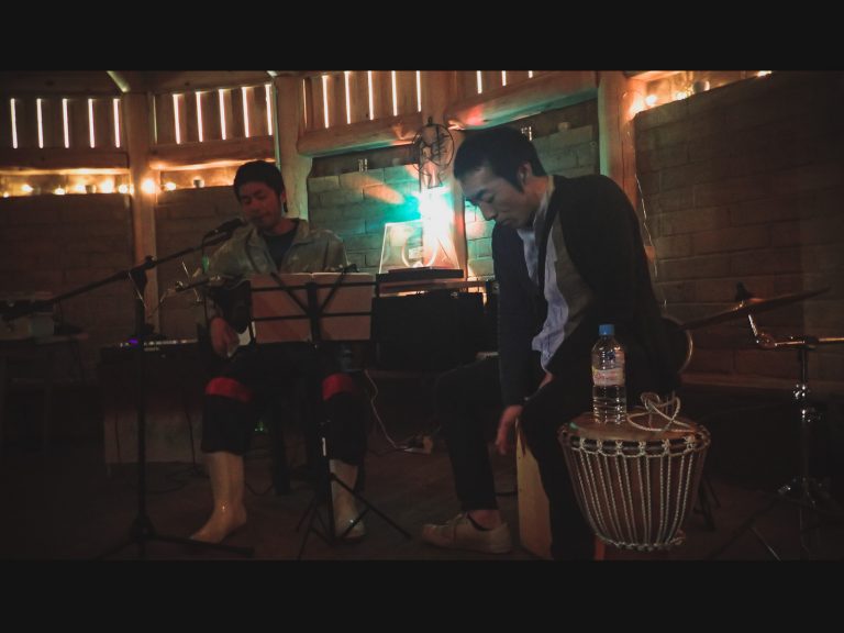 薄暗い木造の屋内で、左側の男性がギターを弾きながら歌い、右側の男性が打楽器のカホンに座って演奏している、静かな雰囲気のライブ演奏の写真