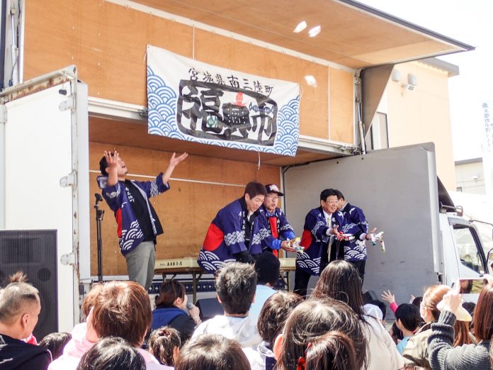 「宮城県南三陸町の復興市」の横断幕が掲げられたトラックの荷台のステージから、法被姿の5人の男性たちが集まった大勢の観客に向けて楽しそうにお菓子を撒いている写真