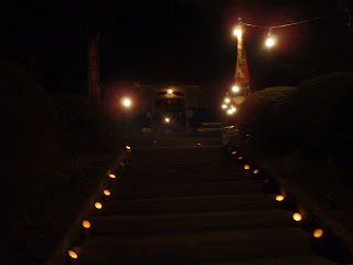 夜の暗がりの中、石段の両脇に並べられた小さな灯りが参道を照らし、その奥には灯りに浮かび上がる神社の社殿が見え、提灯や旗も飾られている写真