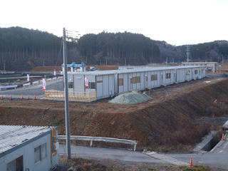 木々が生えた森を背景に、周囲に工事用のコーンやバリケードが設置されている敷地内に、窓が並ぶプレハブ風の長い平屋建物が道沿いに建っている様子の写真