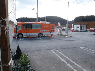 木が生い茂っている林を背景に、周囲を三角コーンに囲まれた駐車場に「ポスクル 移動郵便局」と書かれたオレンジと白の車両が停まっている様子の写真