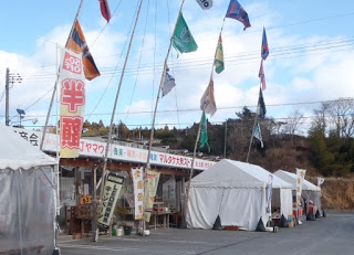 青空の下木々を背景に、「半額」などののぼり旗が掲げられている複数のテントと旗が並ぶ屋外の市場の写真