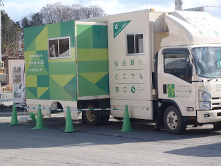 周囲に緑色の三角コーンが置かれている「仙台銀行 どこでも窓口」と書かれた緑と白の移動型銀行トラックが停まっている写真