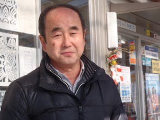 ガラス窓と扉にポスターや飾りが貼られた店舗の前に立っている、黒いジャケットと襟付きシャツを着た商店街の組合長、高橋さんをアップで撮影した写真