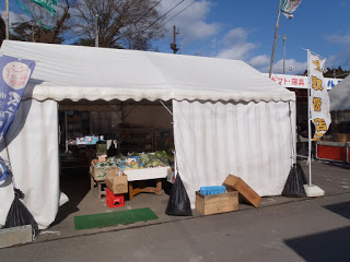 両端にのぼり旗立っている舗装された地面の上に白いテントが立ち、野菜などの商品がテーブルや箱に並べられた仮店舗の写真
