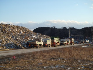 広い敷地の公立志津川病院跡地に、地震や津波で発生したがれきが高く積み上げられ、その周囲の道路には廃棄物を運搬するダンプカーが何台も列をなして停まっている様子の写真