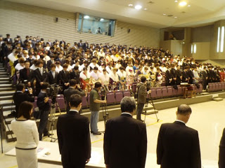 大勢の新成人の方がが階段状の座席に座って前方を見つめており、前方には数名のスーツ姿の人物が立ち、複数のカメラが設置されている式典の会場の写真