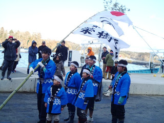 一人の男の子が大漁旗を担ぎ、青いおそろいの法被を着た子どもたちが「寄木ささよ」の伝統行事に参加している様子を写した写真