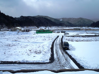 手前には雪に覆われた真っ白な農地が広がり、その中を一本の道路がまっすぐに伸びており、奥には山々が連なって見える冬の風景写真