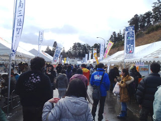 両側にのぼり旗が立てられた出店のテントが並び、通りをたくさんのお客さんが歩いている復興市の賑わいの様子を写した写真